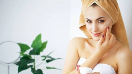 A beautiful young woman in the bathroom taking organic face cream from a jar and applying the cream to the left cheek of her face.