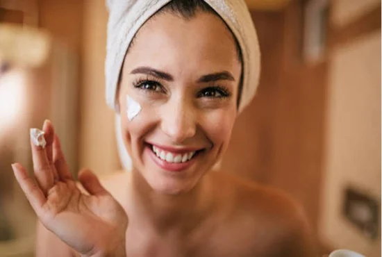 A woman with a smile in the bathroom displaying natural face cream on her finger which she applies above the right cheek of her face.